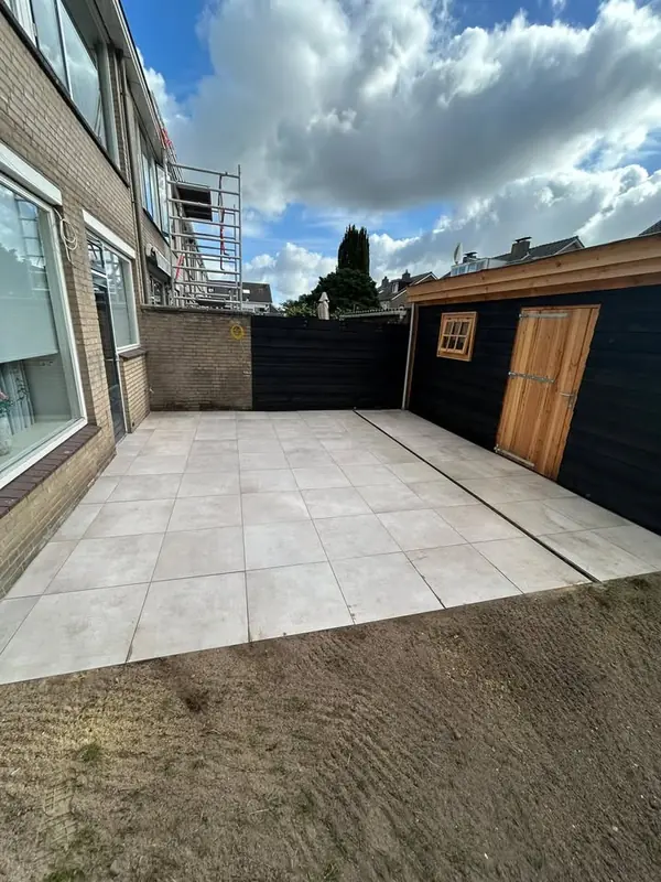 Terras van lichte tegels langs de woning en een zwarte tuinschuur