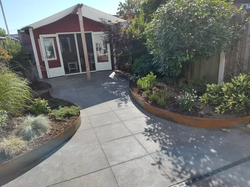 Terras van grote grijze tegels tussen golvende borders met een rand van cortenstaal, met een rood tuinhuis op de achtergrond